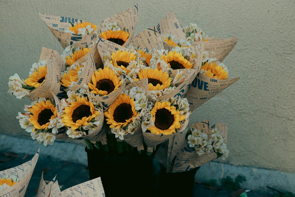 A bouquet of fresh sunflowers with bright yellow petals and dark centers, arranged with smaller white flowers and wrapped in decorative newspaper with printed blue and black text, placed in a black co
