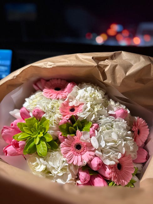 A close-up of a floral bouquet arranged in brown wrapping paper, featuring a combination of pink gerbera daisies, white hydrangeas, and bright green foliage. The flowers are fresh with vibrant petals 
