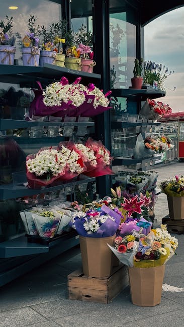 A display of various floral arrangements and bouquets outside a flower shop, featuring fresh flowers such as white chrysanthemums, pink lilies, and purple asters, wrapped in colorful paper, plastic, a