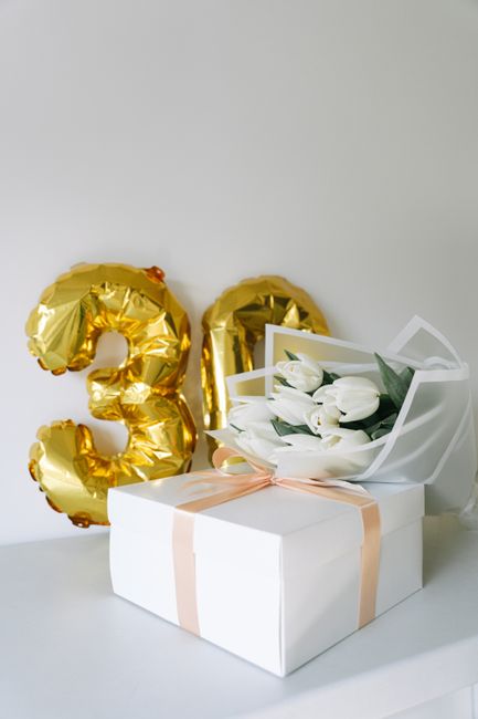 A white gift box with a soft pink satin ribbon tied around it, topped with a small arrangement of fresh white tulips and green foliage in a white paper sleeve. Behind the box, two gold foil balloons s