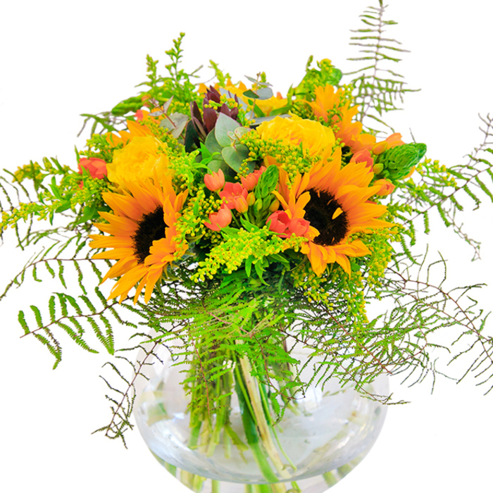 First impressions of this Peckham-inspired bouquet are pure, glowing exuberance-a jubilant mix of sunflowers and buttery yellow roses radiates from a crystal-clear, spherical vase. The sunflowers stand tall, their broad faces open wide, petals in layered gold and ochre, with signature dark, textured centres reminiscent of sunlit afternoons spent in local parks. Threaded among them, spiral yellow roses unfold with a creamy elegance, their scent mingling with the earthy undertones of fresh-cut foliage. Sprays of ruby red hypericum berries shine between the blooms, offering striking punctuation that mirrors Peckham's lively rhythms and colourful art scene. Feathery ferns and airy sprigs of goldenrod spill outwards, softening the lines and suggesting carefree movement inspired by the railway arches' urban greenery. Bluish eucalyptus leaves peek through, providing a cool, sophisticated accent and enhancing the bouquet's harmonious balance. The transparent vase reveals each sturdy stem anchored in clear water, a detail that adds depth and showcases the florist's attention to freshness and form. This bouquet's tactile beauty unfolds with each glance: matte sunflower petals, velvety rose spirals, and the crisp snap of lush greens. Enlivening celebrations from birthdays to spontaneous gatherings, this arrangement expresses joy, togetherness, and a deep-rooted sense of Peckham community-a truly special creation for any moment worth commemorating.